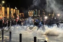 Große Menschenmenge bei Nacht, viele Flaggen, Rauch steigt auf, einige Personen tragen Masken, Lichtquellen im Hintergrund.