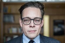 Person mit Brille blickt frontal in die Kamera, Hintergrund unscharfes Bücherregal.