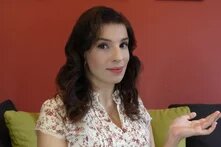 Person sitzt vor roter Wand auf Sofa und hält eine Hand fragend zur Seite hoch.