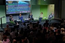 Fünf Personen auf Podium mit Mikrofonen und Notizen, Publikum sitzt davor, im Hintergrund Projektion mit Namen und Titel.