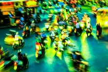 Dozens of people on motorbikes move through a brightly lit, busy street at night, with light trails creating a blurred effect.