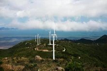 Auf einem grünen Hügel stehen mehrere Windräder, dahinter das Meer und ein bewölkter Himmel. Ein Weg schlängelt sich durch das Gelände.