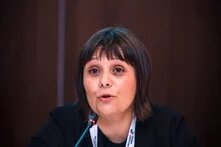 A person with straight, dark chin-length hair speaks into a microphone, wearing a dark top and a white lanyard, seated behind a glass of water.