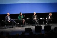 Vier Personen sitzen auf einem Podium, im Hintergrund scheint blau-weiß eine Leinwand. 