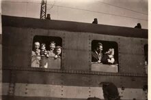 Mehrere Personen lehnen aus den Fenstern eines stehenden Zuges, teils lachend, im Vordergrund eine Person mit Mütze.