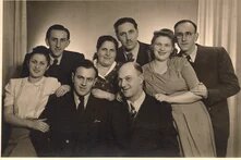 Acht Personen posieren eng beieinander für ein Gruppenporträt vor einem neutralen Hintergrund, mehrere lächeln in die Kamera.