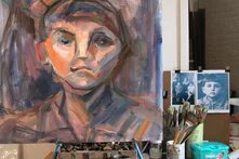 Atelier mit einem Porträtgemälde auf Staffelei, daneben Pinsel, Farbtöpfe, Fotos und Malutensilien auf einem Tisch.