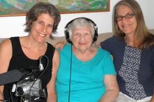 Drei lächelnde Personen sitzen auf einem Sofa, die mittlere trägt Kopfhörer, links vorn steht eine Kamera mit Mikrofon.