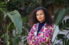 Person mit lockigen, dunklen Haaren, trägt ein pink-gelb gemustertes Oberteil und steht vor einer weißen Mauer mit Grünpflanzen.