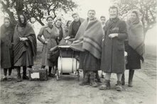 Gruppe von etwa zehn Personen im Mantel oder mit Decke, steht draußen um Kinderwagen, Koffer am Boden, Bäume im Hintergrund.