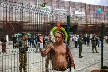 Person with traditional feather headdress andn tatoos stands shirtless in front of a fence; behind the fence are military and people near “COP30 BRASIL” building.