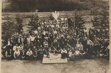 Große Menschengruppe sitzt und steht im Freien, vorne ein Schild mit Noten und Schrift, im Hintergrund Bäume und Zaun. Das Foto ist schwarz-weiß und wirkt alt. Es hat sichtbare Gebrauchsspuren und einen Sepia-Ton.