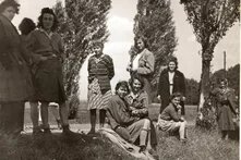 Gruppe von etwa zehn Personen steht oder sitzt auf Wiese mit Bäumen, teils lachend, Kleidung im Stil der 1940er Jahre.