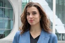 Person mit langen Haaren steht vor einem modernen Gebäude und blickt leicht lächelnd in die Kamera.