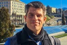 Ein Mann steht draußen vor städtischer Kulisse mit Fahne und Treppen im Hintergrund.