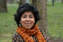 Person with curly dark hair sits outside in front of trees, smiling at the camera.
