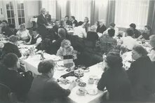 Zahlreiche Personen sitzen an gedeckten Tischen, eine Frau steht sprechend am Rand, auf den Tischen stehen kleine Flaggen.