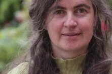 Person mit langen, gewellten Haaren steht draußen, im Hintergrund unscharfe Blumen und Grün.