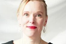 Eine Frau mit zurückgebundenem Haar blickt direkt in die Kamera, Hintergrund ist hell und weich beleuchtet.