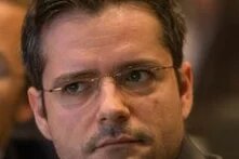 Ein Mann mit Brille schaut ernst zur Seite, im Hintergrund sind verschwommene Personen.