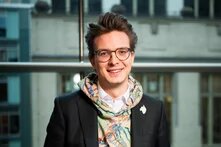 Person mit Brille lächelt in Innenraum vor Fensterfront und blickt in die Kamera.