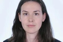 A person with long, dark hair looks neutrally into the camera. She stands in front of a light background, wearing a red top under a dark blazer.