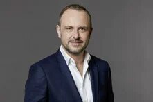 Ein Mann mit kurzem Haar und Bart blickt lächelnd in die Kamera, neutraler Hintergrund.