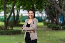 A woman stands in the park with her arms crossed and smiles at the camera, trees and lawn in the background.