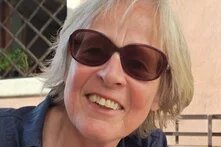 Person mit Sonnenbrille lächelt in Außenbereich, im Hintergrund Fenster mit Gitter und Pflanze.