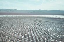 Thousands of mirrors stand in rows in a desert landscape, with low mountains in the background under a lightly clouded sky.