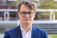 Person mit Brille steht draußen vor einem modernen Gebäude und blickt frontal in die Kamera.