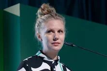 Eine Frau mit Dutt spricht an einem Mikrofon, grüner Hintergrund mit Licht- und Schattenflächen.