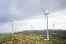 Wind turbines stand on a hilly, green landscape under a cloudy sky, stretching into the distance.