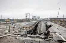 Destroyed road bridge with collapsed center section; guardrail bent, roadway severely damaged, city in the background.
