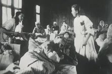 Mehrere Personen liegen dicht an dicht in Betten, teils aufgerichtet, zwei Frauen in heller Kleidung betreuen sie, Raum mit Holzwänden und großen Fenstern.