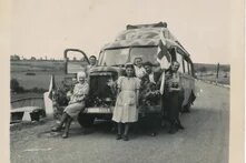 Gruppe von acht Personen vor geschmücktem Bus mit Flaggen und Blumen auf Landstraße, hügelige Landschaft im Hintergrund.