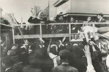 Menschen auf offener LKW-Ladefläche, viele winkende Hände von unten, Häuserfassaden im Hintergrund.