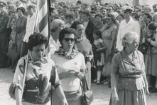 Mehrere Personen mit Halstüchern und Flagge gehen vor großer Menschenmenge, einige in Uniform.