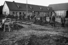 Gruppe gestreift gekleideter Personen gräbt mit Schaufeln im schlammigen Hof vor langem Backsteingebäude mit Scheunen.