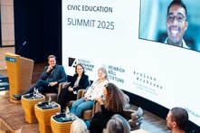 Das Foto zeigt Teilnehmende einer Podiumsdiskussion vor einer Leinwand, auf die ein Video einer weiteren teilnehmenden Person projiziert ist.