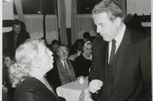 Ein Mann im Anzug spricht ernst mit einer Frau, die ihn ansieht. Im Hintergrund sitzen mehrere Personen an Tischen mit Tassen. Gardinen an den Fenstern.