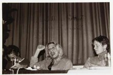 Drei Personen sitzen vor einem Vorhang, mittlere Person hebt die Faust und spricht, die rechte blickt sie an, linke Person hat den Kopf gesenkt.