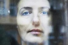 Nahaufnahme eines Gesichts mit blasser Haut und dunklem Haar. Person schaut ernst durch eine leicht reflektierende Scheibe.