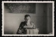 Person sitzt in Uniform hinter Schreibmaschine, Blick nach unten, im Hintergrund Wandteppich mit Pflanzenmotiv.