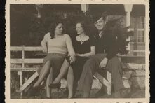 Drei Personen sitzen auf Bank im Freien, zwei schauen sich an, eine Person blickt zur Kamera, Gebäude im Hintergrund.