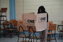 Eine Person steht an einer Wahlkabine mit der Aufschrift „CNE“ in einem leeren Raum mit gestapelten Schulstühlen.