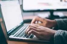 Hände tippen auf Laptop-Tastatur, im Hintergrund ist ein zweiter Monitor mit unscharfem Inhalt zu sehen.