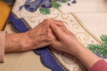 Eine junge Hand hält eine ältere Hand auf bestickter Tischdecke mit weihnachtlichem Muster.