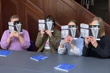 Das Foto zeigt vier Personen, die an einem Tisch sitzen und Flyer einer Veranstaltung vor ihr Gesicht halten.