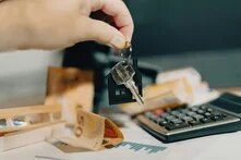 Hand hält Schlüssel mit Haus-Anhänger über Geldscheinen, Taschenrechner und Papieren mit Diagrammen.